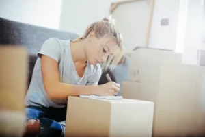 Umzug - Eine Frau sitzt inmitten von Umzugskisten und schreibt etwas auf einen Notizblock der auf eine Kiste liegt.